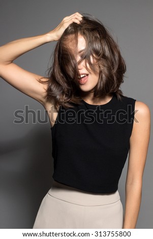 Awesome Caucasian attractive joyful happy sexy female model is shaking head with brunette hair in studio wearing black sleeveless shirt, isolated on gray background