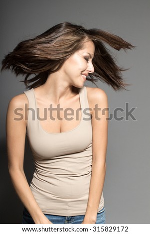 Awesome Caucasian attractive joyful happy sexy female model is shaking head with brunette hair, grimacing in studio, wearing beige sleeveless shirt, isolated on gray background