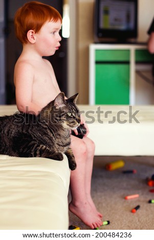 2 year old boy playing a video game while sitting in his living room with his 9 year old, male tabby cat -- image taken in Reno, Nevada, USA