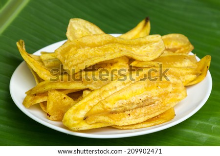 Fried banana chips on white plate with banana leaf background.