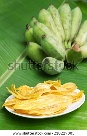 Fried banana chips on white plate with banana leaf background.
