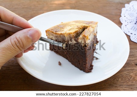 Woman's hand cutting brownie cheese cake by fork on white plate.