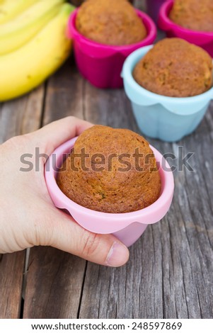 Woman\'s hand holding banana cup cake in colorful baking pan (silicone). On wooden table and banana background.