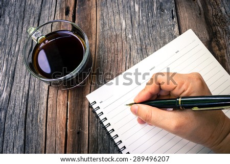 Woman's hand holding a black pen on an empty notebook. Decorate with a glass of black coffee. On wooden table. Hi-contrast and retro style. Dark vignette.