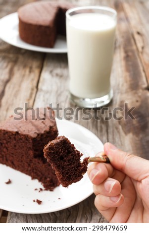 Woman\'s hand picking chocolate cake on white plate. Glass of milk and chocolate cake are background. Over wooden table.