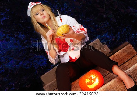 halloween celebration - pretty girl dressed as halloween nurse wearing a blood stained jacket holding a pumpkin and a syringe and a dark forest background.