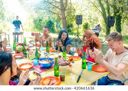 group of friends at garden barbeque lunch smiling at colorful lunch table while cooking at the smoking grill in sunny park background - weekend get-together lunch with family friends