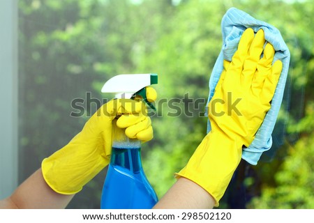 Hands in a yellow glove wiping  window with napkin and the cleaning fluid on the background of trees