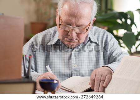 Old man in glasses writing from the books in the room. Close portrait
