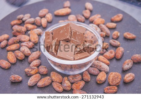 A lot of brown chocolate, in a glass bowl. Near cocoa beans