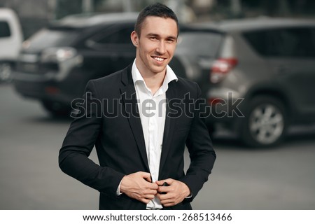 Young successful smiling man in a black suit, on the street, on the background of cars. Happy modern man. Outdoors