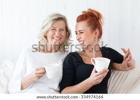 Two beautiful women-a mother and daughter sitting on the sofa at home, snuggled. They drink coffee, tea. Relationships and shared time.