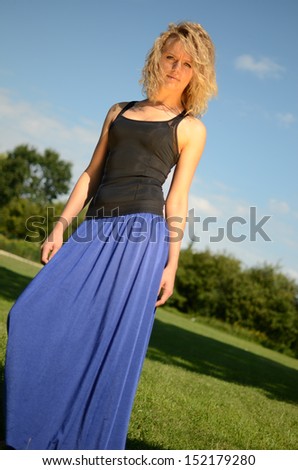Female model with blond, curly hairs. Girl wearing blue dress and black top. Outdoor photo session in the park with blue sky as background.