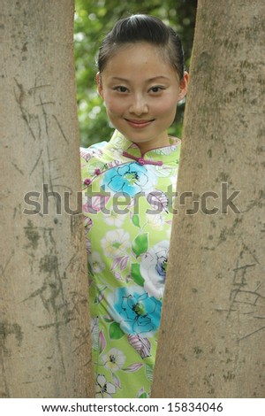 Chinese girl in park. Standing between trees, relaxing with kind smile, wearing traditional Chinese dress.