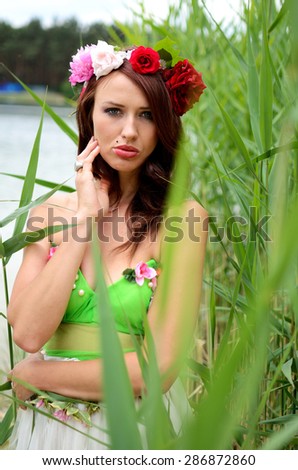 Portrait of female model with chestnut hairs, wearing wreath made of mixed flowers. Woman posing near lake surrounded by reeds.