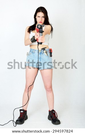 Young female model wearing white top and jeans\' shorts. Girl holding big driller, keeping key, screwdriver and small hammer in her pocket.