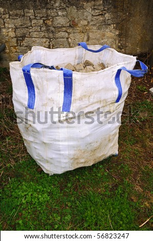 White big bag with blue handles and stones inside in front of an old stone wall