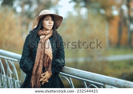 Beautiful brunette girl in a hat and coat standing on the bridge and looks afar
