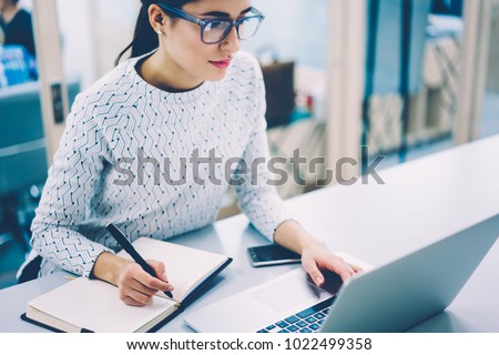 Skilled female web designer in eyewear making online research planning project writing ideas in notepad,administrative assistant booking tickets on web page sitting at working place in office