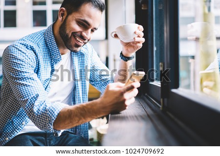 Cheerful male person happy to receive messages from friend checking mail via smartphone during rest in cafe, smiling man satisfied with getting good news using telephone fr networking drinking coffee