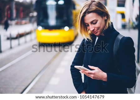 Smiling young woman checking table of city traffic online on web page using smartphone,positive hipster girl satisfied with app for paying for electric transport via cellphone waiting for tram