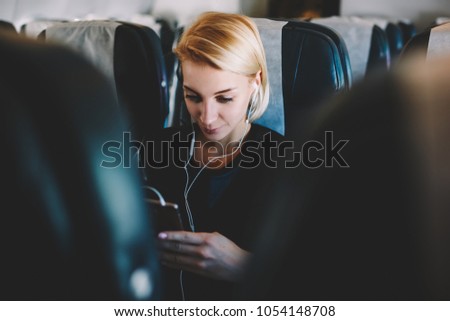 Attractive female passenger of airplane in modern earphones spending time during flight on watching interesting movie online on digital smartphone device satisfied with wireless connection on board