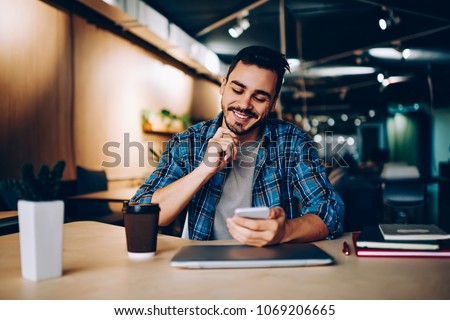 Smiling hipster guy reading new message in social networks confirming new friend on smartphone, positive young man satisfied with banking app on mobile phone checking balance while learning