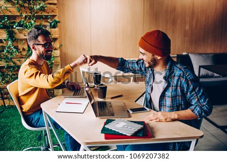 Prosperous male it developers clinking fists in sign of agrement cooperating on common startup project, young hipster guys greeting while meeting in coworking space for working creative together