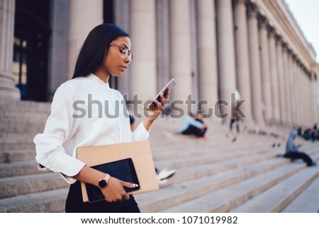 Pensive dark skinned student of faculty of law checking mail and reading notification with financial news on smartphone device connected to smartphone standing outdoors near university building