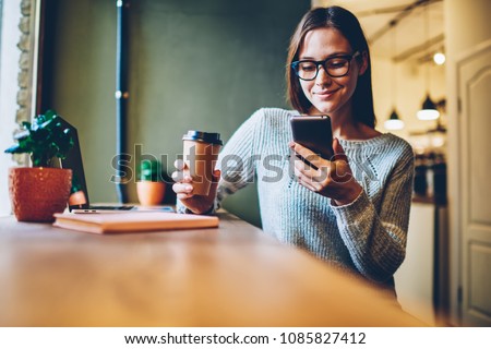 Cheerful hipster blogger with coffee to go in hands reading incoming sms message on smartphone connected to free wifi.Positive young student watching broadcasting online on modern mobile phone