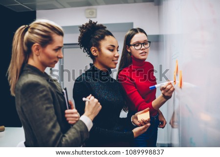Clever female student writing on board her idea for explaining to colleagues, multiracial crew of employees team working on startup using flip chart for planning successful strategy and discussing
