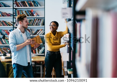 Positive african american female librarian helping caucasian student to search literature book in bookshelf.Cheerful two young people in casual wear discussing new bestseller standing in library