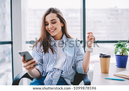Cheerful young woman checking new feed and content in social networks via smartphone during coffee break in office,happy cute female blogger satisfied with followers messages sitting at desktop