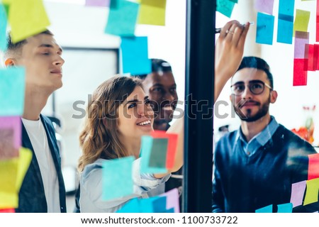 Diverse team of positive young people laughing while working together during brainstorming and standing behind glass wall with sticky colorful papers.Cheerful students learning words from stickers