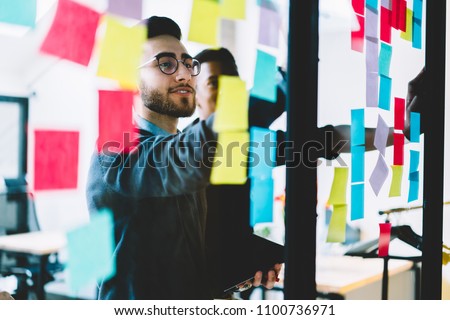 Diverse team of skilled young people learning foreign words from colorful stickers glued on glass wall during collaborative process in office interior.Multicultural students using paper sticky notes