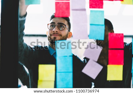 Young creative man together with african american colleague reading foreign words written on colorful stickers for better learning language standing behind glass wall in office interior