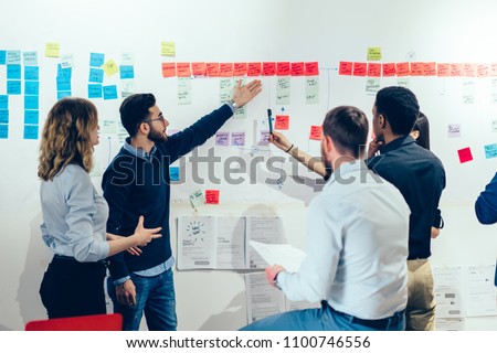 Intelligent male professional pointing on colorful stickers with text message glueded on wall and discussing information with creative multicultural colleagues having brainstorming meeting in office