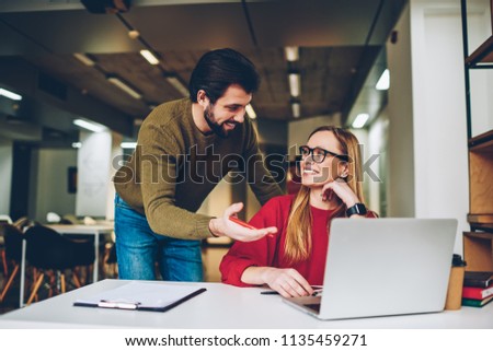 Young work employee in eyeglasses enjoying break while working on laptop computer with male colleague standing by, two smiling hipsters bloggers remember funny video vlog creating together indoors