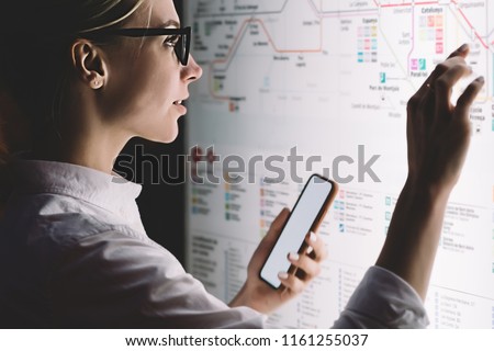 Interactive kiosk with public transport subway map.Female standing at big display with smartphone in hand.Young woman touching with finger screen while using train schedule application on mobile phone