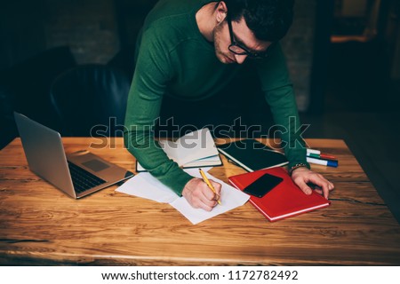 Good looking pensive caucasian employee carefully creating organisation plan during paper work, serious pondering architect writing main theses for startup project standing near wooden desktop