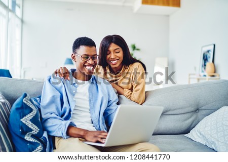 Cheerful happy African American young marriage laughing while watching movie on laptop computer at home interior, happy dark skinned couple in love shopping online on netbook enjoying store discounts