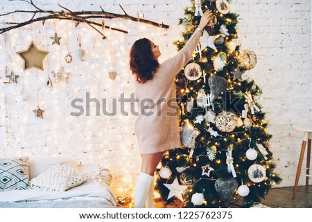 Young female in knitwear decorating traditional Christmas green tree in cozy home interior during vacations, charming woman feeling joy while making design for winter holidays at modern apartment