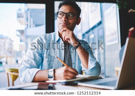 Thoughtful hindu student pondering on creative ideas for writing essay during exam preparation at modern netbook with wireless 4G internet.Contemplative hipster guy in denim jacket drawing sketch