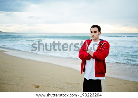 Young athlete man in white t-shirt and sports a red windbreaker standing on the beach back to the sea with his arms crossed over his chest and a serious look