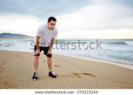 Young  man in white shirt leaning on his knees and breathing hard after a grueling jogging on the beach during a storm