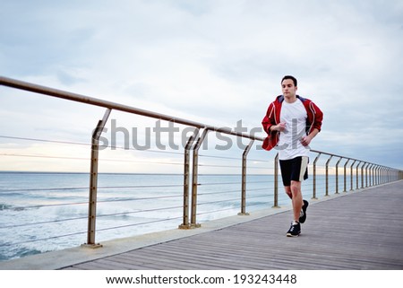 Full length portrait of male runner working out outdoors at early morning, young athlete running ob wooden pier with amazing sea view on background