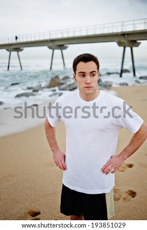 Young athlete man in white t-shirt put both hands on the sides and standing on the beach looking away, portrait athlete resting after jogging