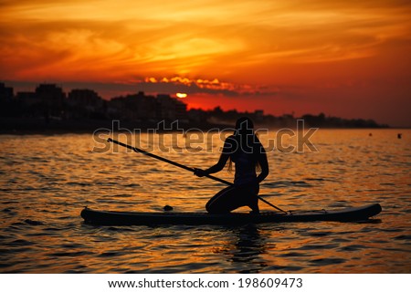 Silhouette of a beautiful girl floating at sea on paddle surf board encountering stunning sunrise