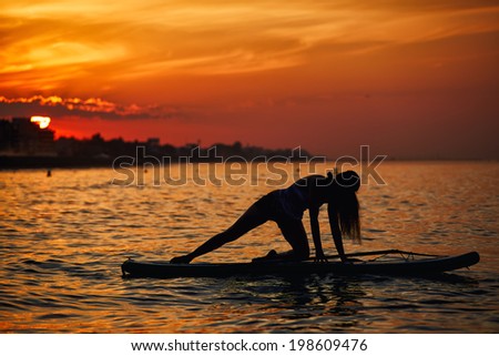 Thin silhouette of girl engage yoga on the paddle board on the bright backboard of the african orange sunset reflected on the ocean