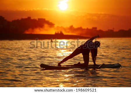 Slim silhouette of girl doing yoga on the paddle board on the bright backboard of the african orange sunset reflected on the ocean